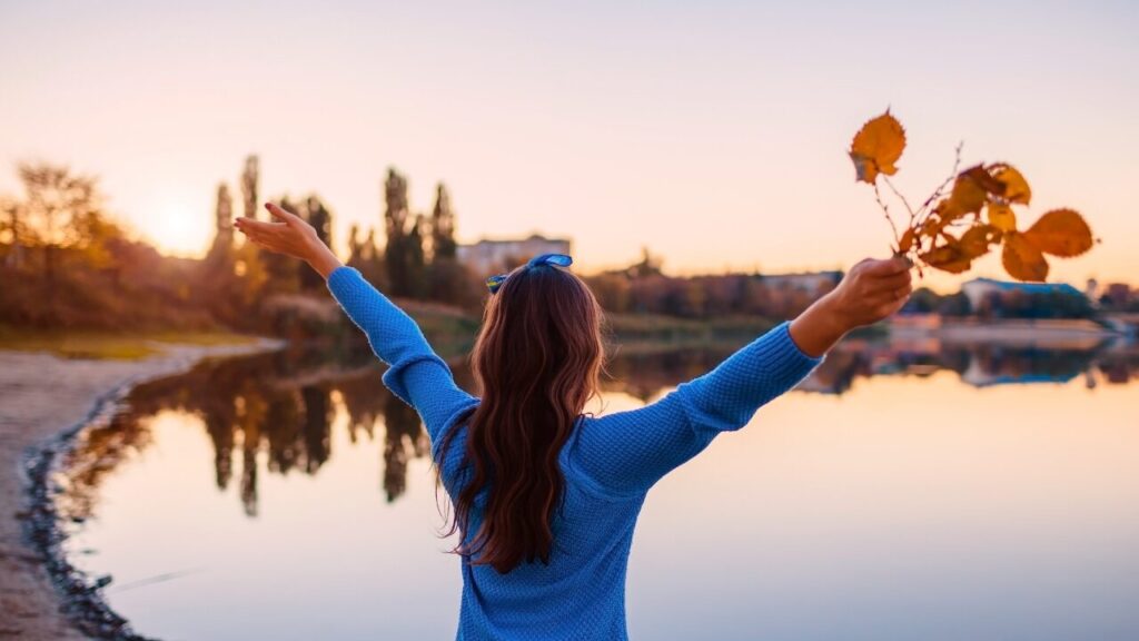 女の人が小枝の束を持ちながら湖に向かって両手を上げてている画像。