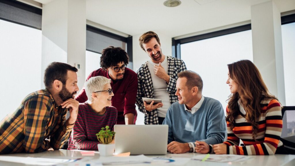 コワーキングスペースを利用して６人の人が楽しそうに談笑している様子。デスクの上にはノートPCやグラフ資料などが置かれている。